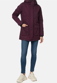 Giacca con cappuccio color bordeaux con due tasche anteriori, chiusura con cerniera e bottoni a pressione; tessuto strutturato; abbinata a jeans blu e scarpe da ginnastica bianche.