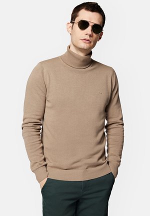 Beżowy sweter z golfem, wykonany z gładkiej dzianiny, o dopasowanym kroju i prążkowanych mankietach. Mały logo na klatce piersiowej, noszony z ciemnymi spodniami.