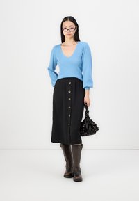 Top de manga longa às riscas em azul claro com decote em V, acompanhado de uma saia midi preta com botões frontais, botas castanhas de pele até ao joelho e uma mala preta.