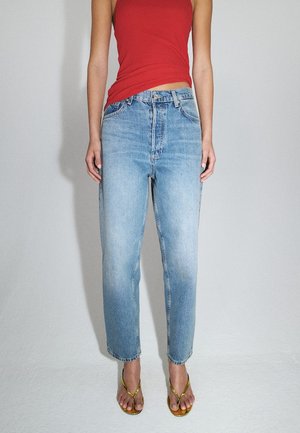 Persoon draagt lichtblauwe high-waisted jeans, rode mouwloze top en gouden sandaaltjes met bandjes, staand tegen een eenvoudige lichte achtergrond.