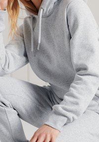 Felpa con cappuccio grigio chiaro con coulisse, polsini a costine e finitura testurizzata, abbinata a pantaloni jogger coordinati, che mostrano un design casual.