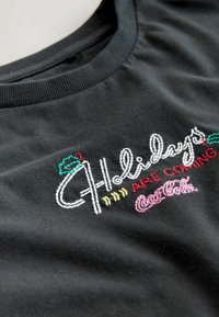 Black cotton t-shirt featuring colorful embroidered text: "Holidays are Coming" with holly and Coca-Cola logo accents.