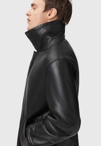 Sort læderjakke med høj krave, knaplukning og tekstureret finish. Designet giver en slank silhuet med sidelommer.