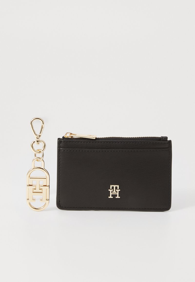 Porte-cartes en cuir noir avec fermeture éclair et quincaillerie dorée. Présente un logo sur le devant et un porte-clés détachable avec un design de logo similaire.