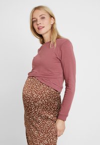 T-shirt de maternidade em rosa suave, com mangas compridas e corte descontraído, combinada com uma saia justa castanha com bolinhas brancas. Tecido texturizado.