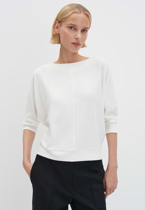 Witte gebreide trui met een brede halslijn, driekwart mouwen en een geribbelde structuur; gecombineerd met een zwarte broek.