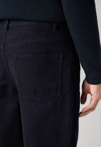 Donkerblauwe broek van duurzaam materiaal, met een enkele achterzak met een rechthoekig, gestikt ontwerp en een textuurdetail aan de zijkanten.