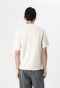 Polo à manches courtes, couleur beige clair, avec un motif texturé, un col côtelé et un tissu doux, associé à un pantalon gris.