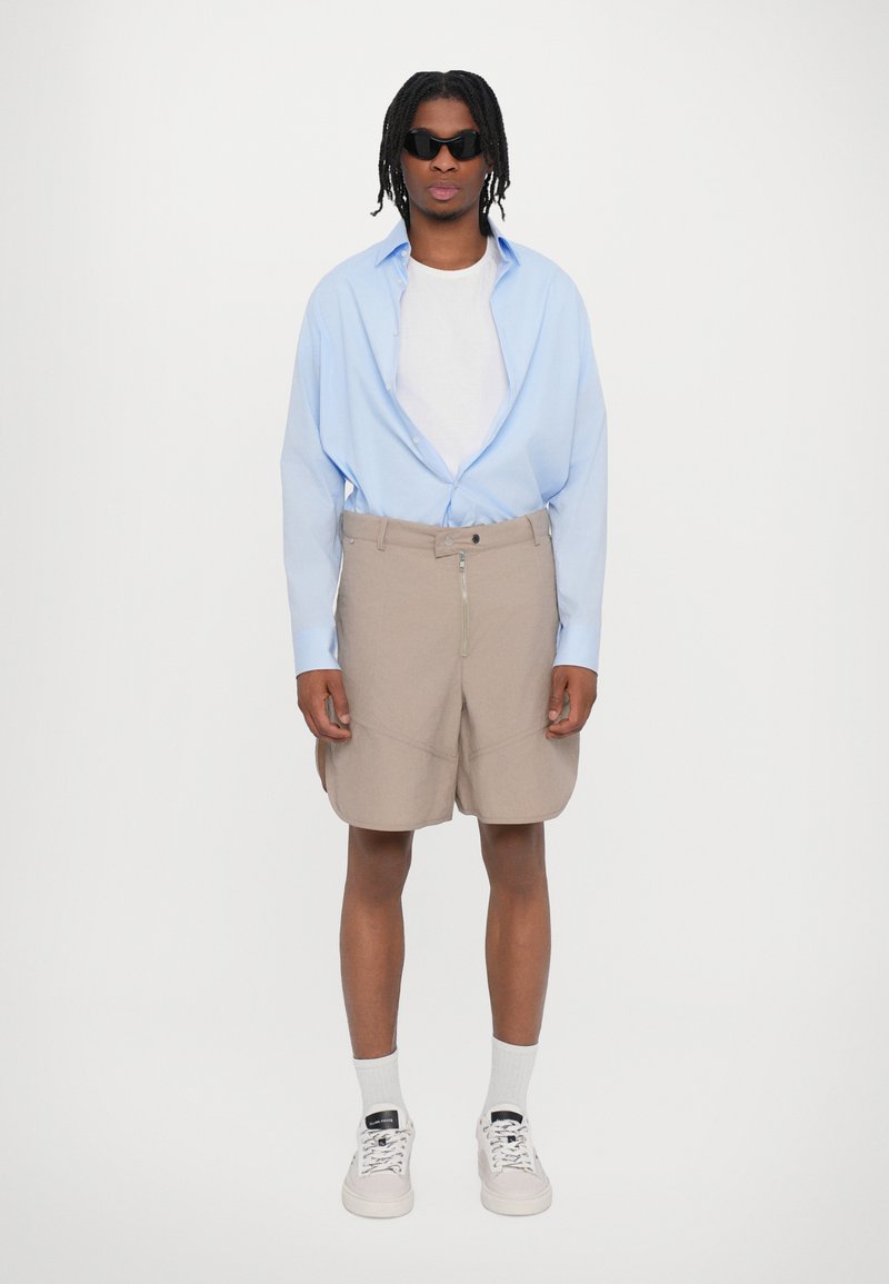 Light blue oversized shirt, white t-shirt, beige shorts with zipper detail, white socks, and light sneakers. Dark sunglasses compliment the outfit.