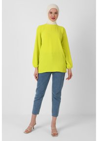 Langærmet, lys gul tunika med høj hals og rynkede manchetter, parret med lyseblå slim-fit jeans og beige sandaler.