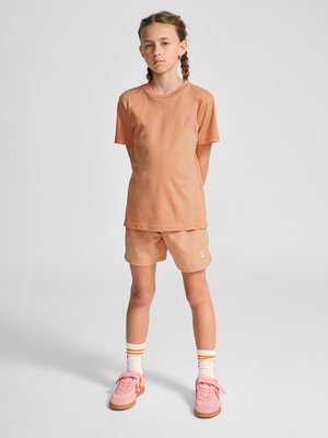 Ung pige med fletninger iført ferskenfarvet t-shirt og shorts, hvide sokker med orange striber og pink sneakers, står med hænderne på ryggen.