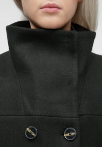 Pogled od blizu na osobu koja nosi tamnosivu kapu s visokim ovratnikom i dva okrugla dugmeta, vidljive usne i donji dio lica.