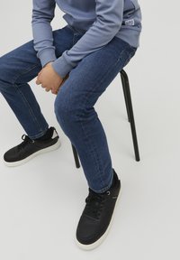 Zilas džinsi un gaiši pelēks sporta krekls, valkāti ar melniem apaviem ar baltu zoli, sēž uz melnas krēsla pretī gaišai fona.