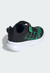 Zapatilla deportiva negra con patrones geométricos verdes, una tira de velcro y un contrafuerte texturizado en el talón sobre una suela blanca.