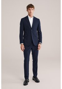 Mężczyzna ubrany w dopasowany granatowy garnitur, białą koszulę i czarne skórzane mokasyny, stojący na tle jednolitego beżowego tła.