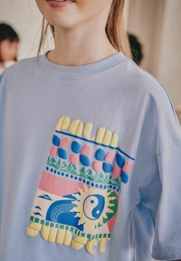 Ljusblå bomulls t-shirt med en färgglad grafisk design med "Palm Sunset", inklusive blomster- och solmotiv i livfulla nyanser.