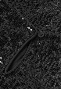 Gros plan sur un tissu en sequins noirs avec un rabat à boutons et une ouverture, montrant une texture brillante et des coutures fines.