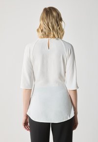 Haut blanc à manches trois-quarts, avec une texture subtile et un détail en trou de serrure au dos. Porté avec un pantalon noir.