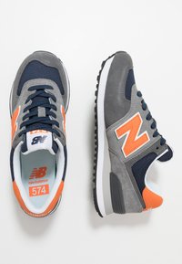 Sneakerii New Balance 574 au o parte superioară din suede și plasă gri, accente bleumarin, un logo portocaliu și o talpă din cauciuc negru cu o talpă intermediară albă.