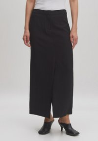 Jupe noire longue jusqu'au sol en tissu texturé. Présente une coupe droite avec une fente sur le côté. Accompagnée de mules aiguës noires à talons.