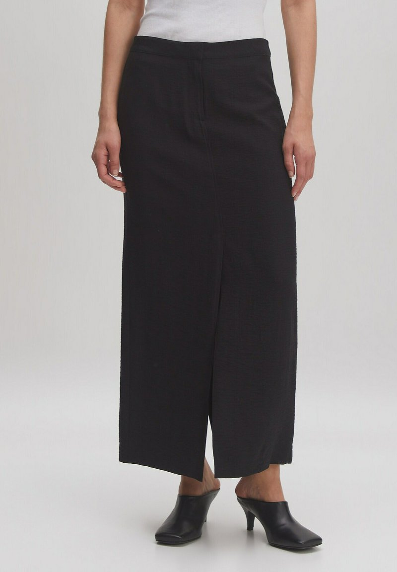 Jupe noire longue jusqu'au sol en tissu texturé. Présente une coupe droite avec une fente sur le côté. Accompagnée de mules aiguës noires à talons.