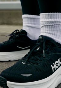 Adidași negri cu talpă și logo alb, purtați cu șosete albe cu dungi pe asfalt.