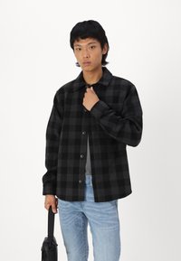 Veste à carreaux noire et grise, à boutons, à manches longues, en mélange de laine, portée par-dessus une chemise grise avec un jean bleu clair.