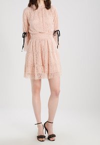 Spetklänning i mjuk rosa med strukturerat mönster, puffärmar och svarta knytdetaljer vid ärmsluten. U-formad kjol med en figurnära midja. Svarta klacksandaler.