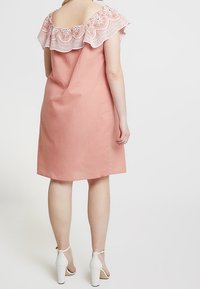 Persikka mekko, jossa on röyhelöillä varustettu pitsikaulus, polvipituinen leikkaus ja takakuvio; yhdistettynä valkoisiin nilkkaremmikenkiin. Sujuva tekstuuri.