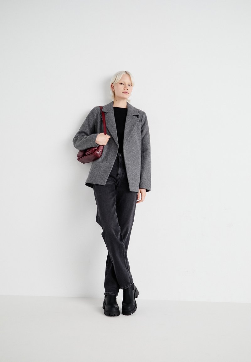 Blazer grigio con texture, top nero, jeans scuri e stivaletti neri; la modella tiene una piccola borsa bordeaux. Design minimalista su uno sfondo chiaro.