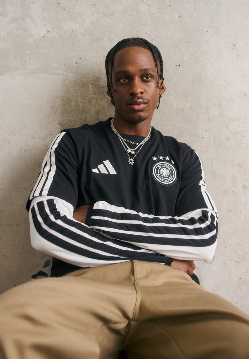 Νεαρός άνδρας με πλεγμένα μαλλιά, φορώντας ένα μαύρο �και άσπρο μακρύ μανίκι μπλούζα Adidas με λογότυπο γερμανικής ποδοσφαιρικής ομάδας, καθισμένος μπροστά σε έναν τσιμεντένιο τοίχο.