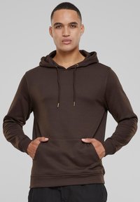 Felpa con cappuccio marrone, dotata di tasca frontale, dettagli con stringhe e polsini a coste. Caratterizzata da una texture morbida e un design casual.