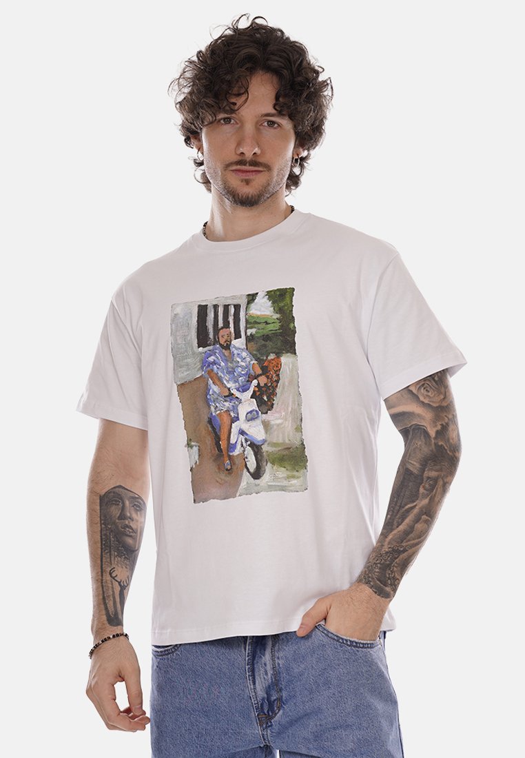 Uomo con capelli ricci e tatuaggi che indossa una maglietta bianca con una grafica colorata di una persona su uno scooter, abbinata a jeans blu.