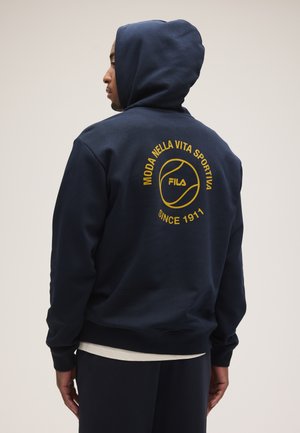 Mężczyzna noszący granatową bluzę z kapturem z żółtym logo FILA i włoskim tekstem "Moda Nella Vita Sportiva Since 1911" na plecach, zwrócony w lewo.