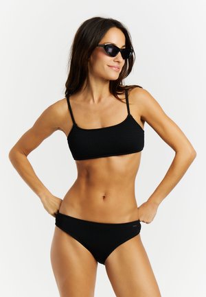 Femme portant un bikini noir texturé et des lunettes de soleil noires à monture œil de chat, posant les mains sur les hanches devant un fond blanc uni.