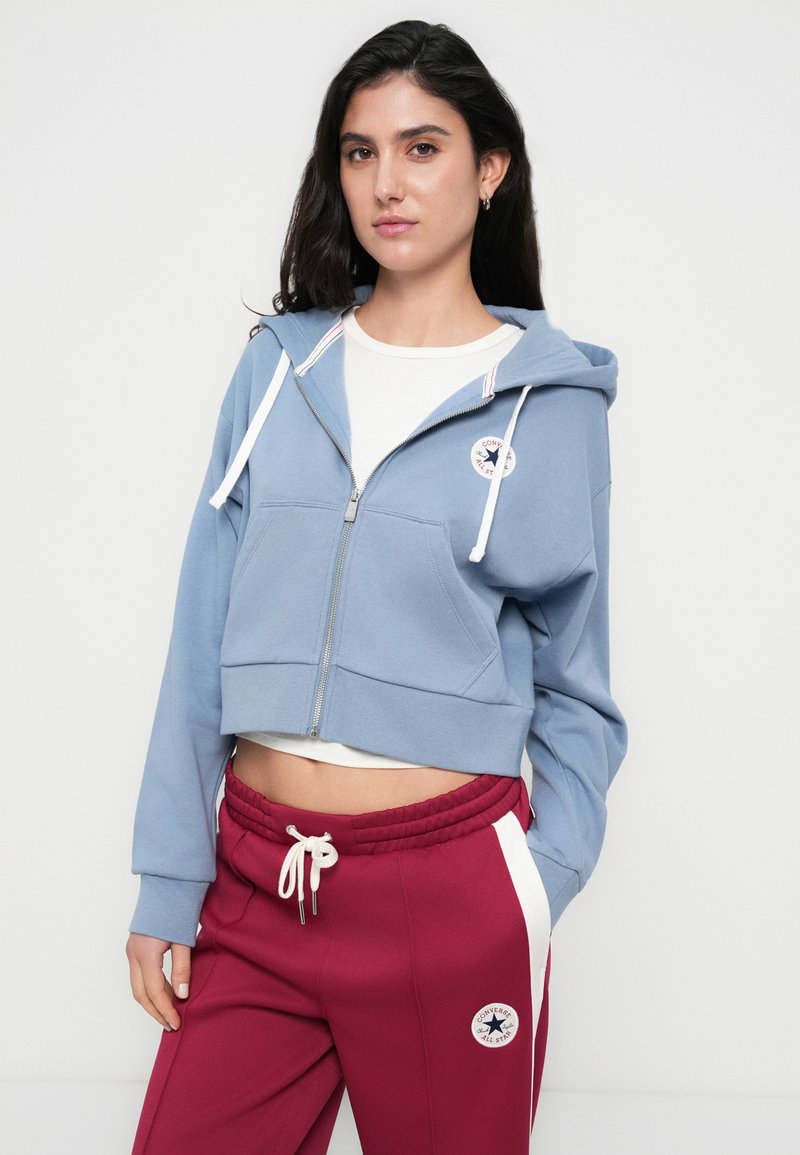 Klipset blå hættetrøje med lynlås foran, hvide snore og en logo-patch. Kombineret med vinrøde bukser med en hvid sideliste.