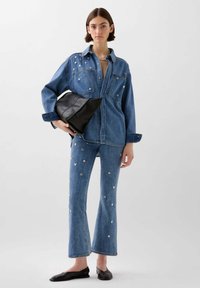 Giacca di denim e jeans a zampa d'elefante con applicazioni a forma di cuore. Una modella tiene in mano una borsa a pochette in pelle nera, in piedi davanti a uno sfondo neutro.