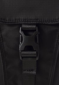 Bolsa de tela negra con una superficie texturizada, que cuenta con una hebilla y una correa negras. El diseño incluye patrones en cuadrícula y detalles de costura reforzada.