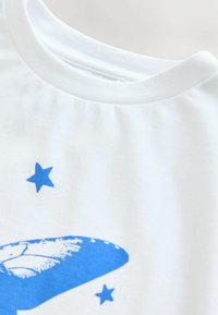 Biele bavlnené tričko s okrúhlym výstrihom, zdobené modrou grafickou grafikou motýľa a modrými hviezdami. Tkanina vyzerá hladko a je mierne štruktúrovaná.