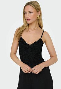 Camisole noir avec des détails en dentelle et des bords en forme de vagues. Dotée de fines bretelles, portée avec des bijoux en or, y compris un collier et des boucles d'oreilles.