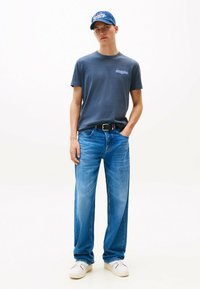 Camiseta de algodón azul con un pequeño logo, combinada con jeans rectos de color azul claro. Accesorios incluyen una gorra azul y zapatos blancos.