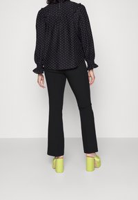 Zwarte blouse met witte stippen en ruches, gecombineerd met zwarte flared broek en groene plateauhakken. Achteraanzicht weergegeven.