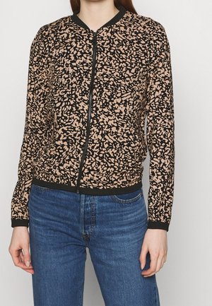 Personne portant une veste zippée beige et noire à motif abstrait associée à un jean bleu sur un fond uni.