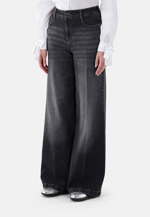 Woman wearing high-waist wide-leg faded black jeans with front pockets, paired with a white blouse with tied cuffs and silver shoes.