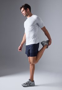 Tricou sportiv gri deschis, pantaloni scurți navy închis și pantofi de alergare gri. Bărbat întinzând o picior cu piciorul ridicat, în interior.