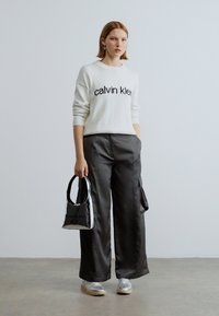 Biely pletený sveter s čiernym nápisom "calvin klein", skombinovaný s tmavosivými nohavicami so širokými nohavicami, držiaci malú čiernu kabelku, obutý v strieborných teniskách.