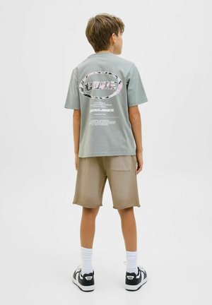 Grå t-shirt med kort ärm och grafiskt tryck på ryggen, i kombination med beige shorts. Modellen bär vita strumpor och svarta sneakers.