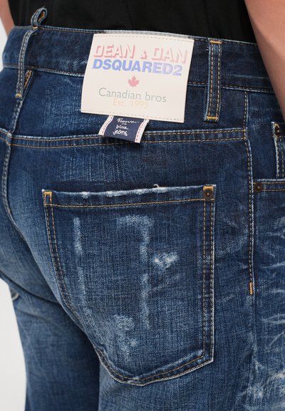 Prim-plan al unui buzunar din spate al unor blugi din denim albastru, cu detalii uzate și o etichetă albă pe care scrie „Dean & Dan DSQUARED2 Canadian bros Est. 1995”.