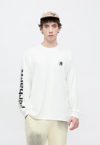 Langærmet hvid T-shirt med lille sort logo på brystet og fed skrift ned langs venstre ærme. Båret med beige bukser og en tan-kasket.