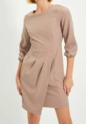 Femme portant une robe taupe mi-longue avec des manches trois-quarts bouffantes et un détail de jupe plissée asymétrique sur fond blanc.